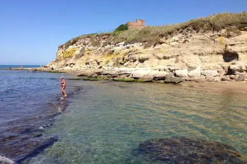 Une femme en bikini profite de la mer limpide près d'une falaise rocheuse à Anzio, avec une tour ancienne en arrière-plan.