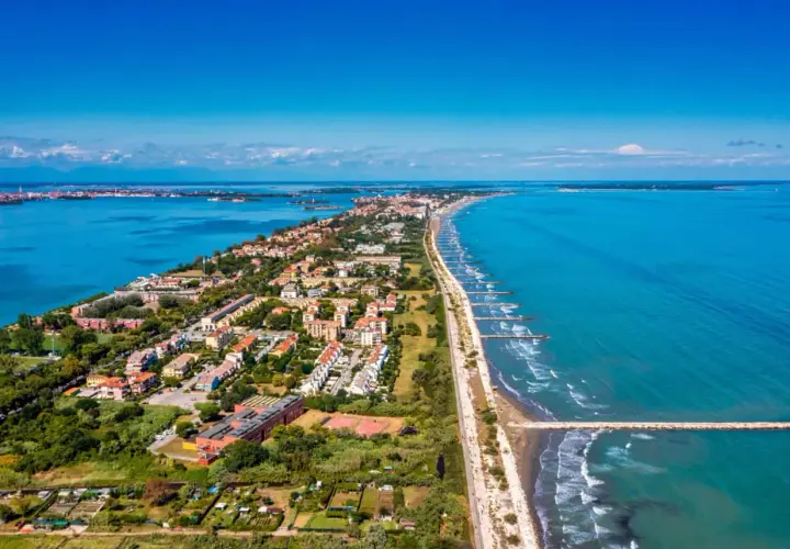 Vue aérienne du Lido di Venezia, une longue bande de terre bordée par la mer Adriatique et la lagune, avec des maisons, des arbres et des digues.