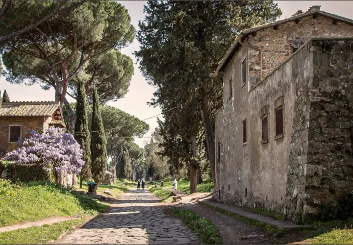 Chemin pavé bordé de cyprès et de pins, avec des maisons anciennes et des glycines en fleurs. Un air de via antica Roma.