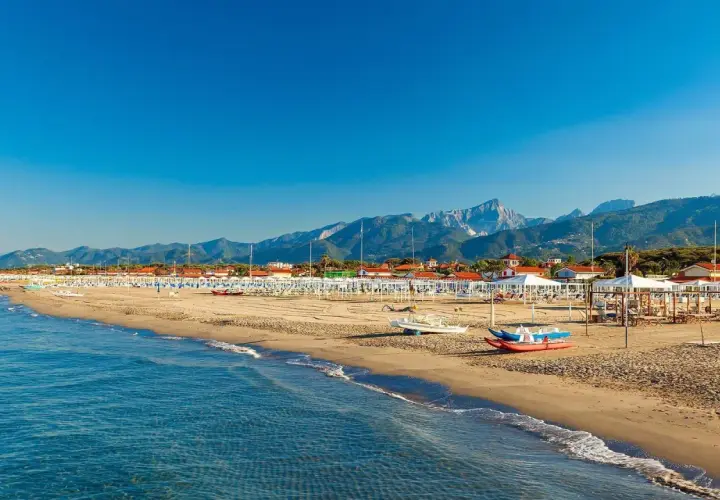 Plage de sable fin à Forte dei Marmi, Italie, avec des rangées de parasols blancs et des montagnes majestueuses en arrière-plan.