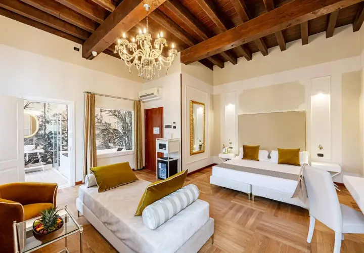 Suite luxueuse au **hotel Cariola Caprino Veronese** avec lit king-size, canapé, lustre et vue sur jardin tropical.