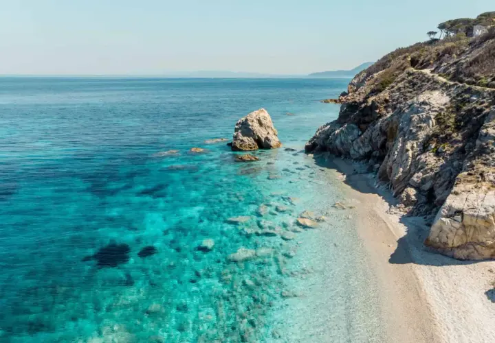 Plage de sable fin bordée par des eaux turquoise cristallines et des falaises rocheuses, typique des iles toscanes. Un rocher solitaire émerge de la mer.