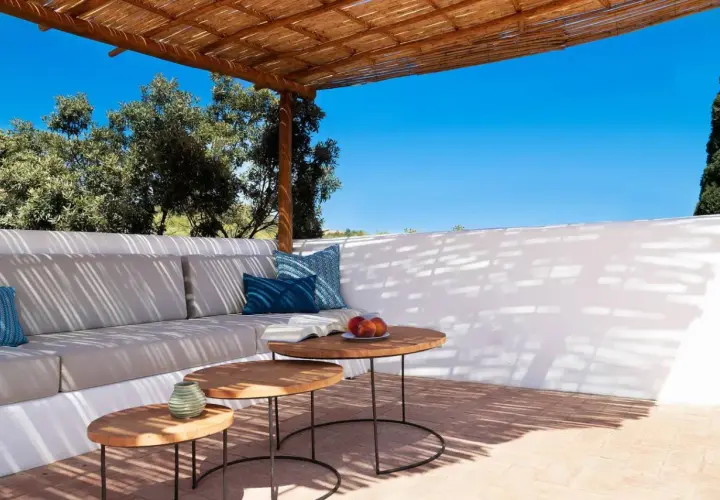 Terrasse ombragée d'un agriturismo en Sardaigne, avec canapé confortable, tables basses en bois et vue sur la végétation luxuriante sous un ciel bleu éclatant.