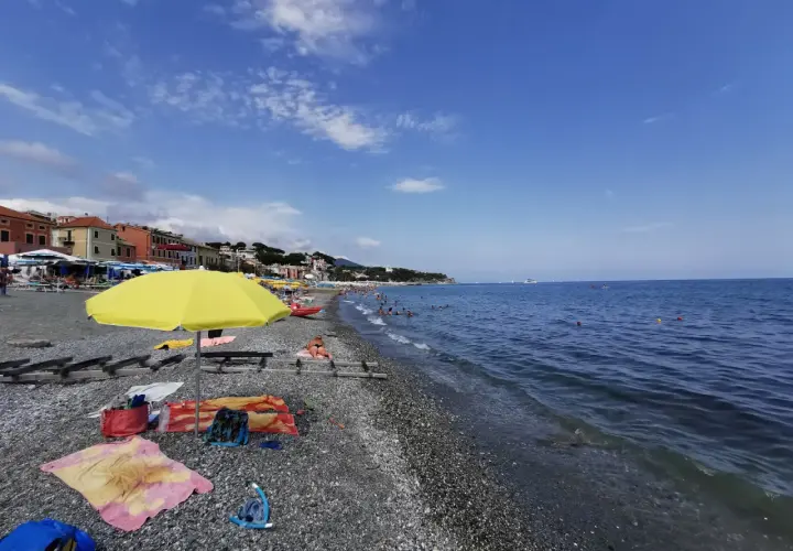 Plage de galets animée, avec des parasols, des serviettes et des baigneurs profitant du soleil. La mer bleue s'étend à l'horizon, avec des bateaux au loin. Un aperçu de la vie sur la côte ligure.