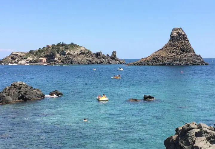 Vue sur la mer bleue et les rochers volcaniques d'Aci Trezza, Sicile. Des bateaux et des baigneurs profitent du soleil.
