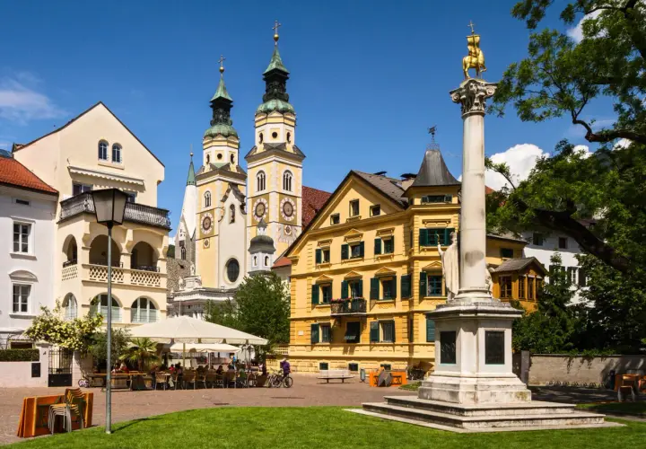 Place ensoleillée à Bressanone avec des bâtiments colorés, une colonne monumentale et des terrasses de café animées.