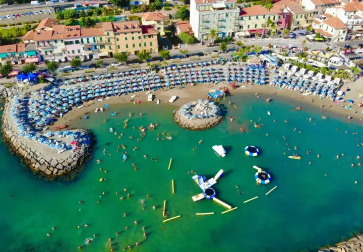 Plage animée à Arma di Taggia, Italie, avec des parasols bleus et blancs, des baigneurs dans la mer turquoise et des structures gonflables.