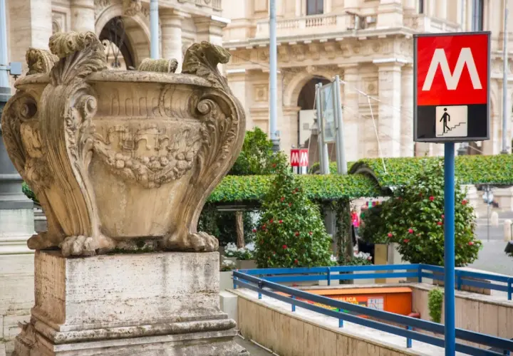 Un signe du **metro vatican** indique l'entrée d'une station, à côté d'une urne antique et de verdure luxuriante.