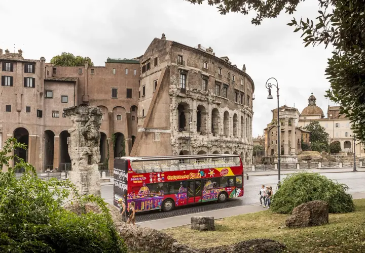Un bus touristique rouge vif, "Citysightseeing Roma", passe devant des ruines antiques et des bâtiments romains.