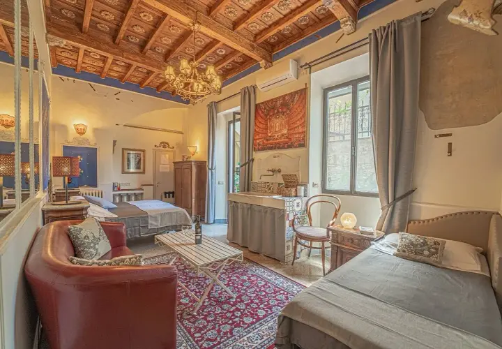 Chambre d'hôte toscane avec lits jumeaux, canapé rouge, tapis persan et plafond en bois sculpté.