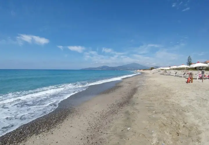 Plage de sable et de galets à Ascea Marina, avec des vagues bleues, des parasols blancs et des montagnes à l'horizon.