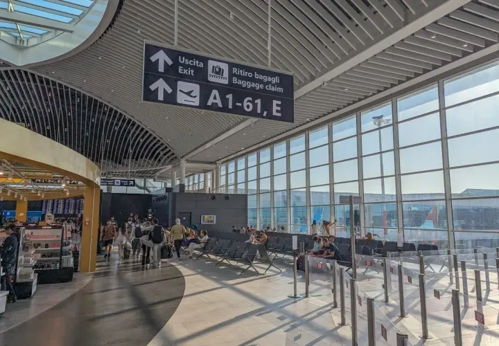 Intérieur lumineux d'un aéroport, avec des panneaux indiquant "Uscita" et "Ritiro bagagli". Des voyageurs attendent près des grandes baies vitrées, prêts pour leur vol au départ de l'aéroport Rome centre ville.