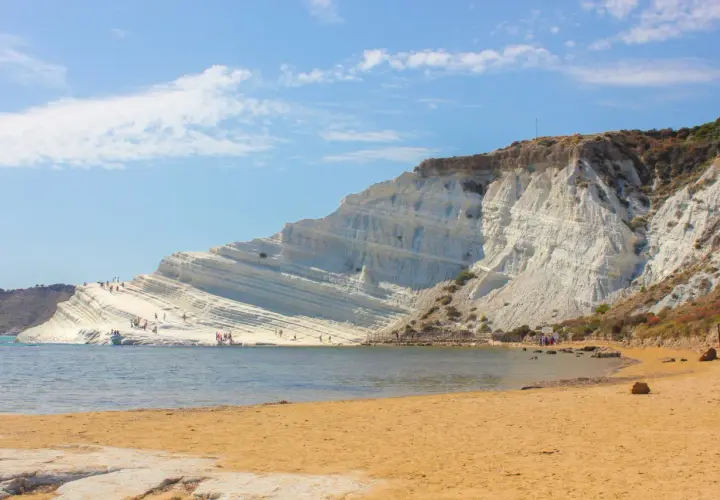 La Scala dei Turchi, une falaise blanche spectaculaire en Sicile, se dresse au-dessus d'une plage de sable doré et d'une mer bleue. Des visiteurs explorent ses terrasses naturelles.