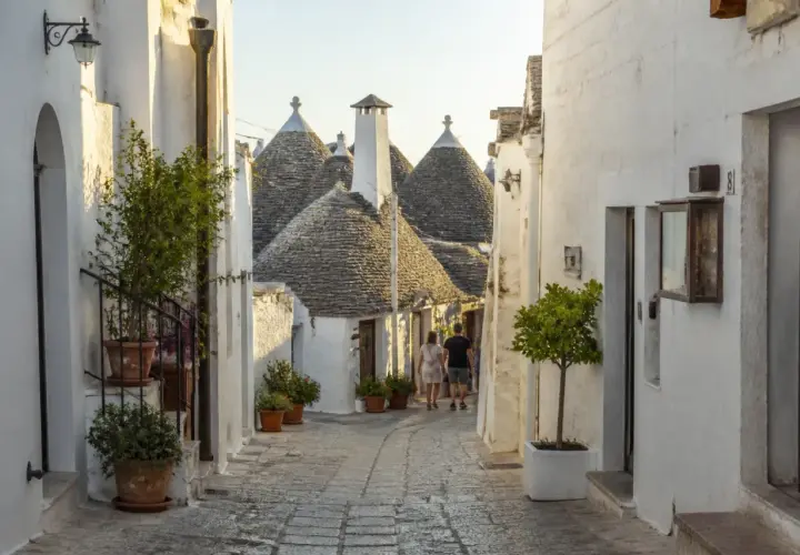 Ruelle pavée bordée de maisons blanches aux toits coniques typiques des Pouilles à éviter, avec un couple se promenant.
