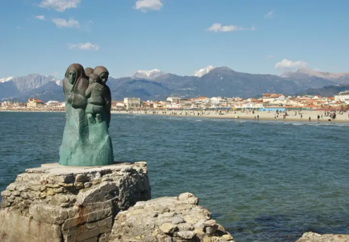 Sculpture de famille sur un rocher, face à la mer et à la ville de via Reggio, avec des montagnes enneigées en arrière-plan.