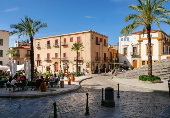 Place animée de Cefalù avec des terrasses de café, des palmiers et des bâtiments historiques sous un ciel bleu.