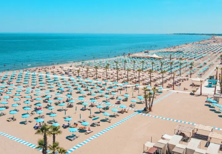 Plage de Chioggia, Italie, avec des rangées infinies de parasols bleus et blancs, des chaises longues et des palmiers sous un ciel bleu éclatant.