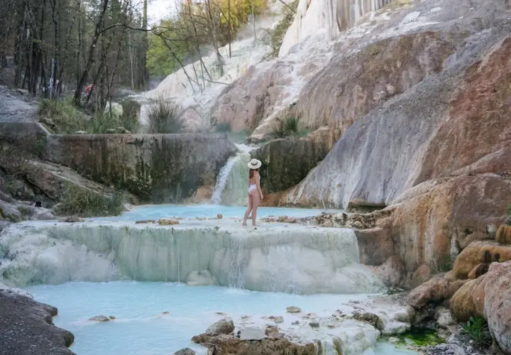 Une femme en maillot de bain blanc et chapeau de paille admire les cascades et les bassins turquoise des Bagni San Filippo, un paysage naturel spectaculaire.