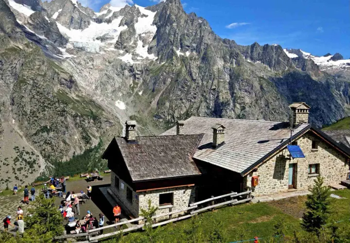 Refuge de montagne en pierre, idéal pour une location vacances Aoste Italie, avec des randonneurs profitant du paysage alpin spectaculaire.