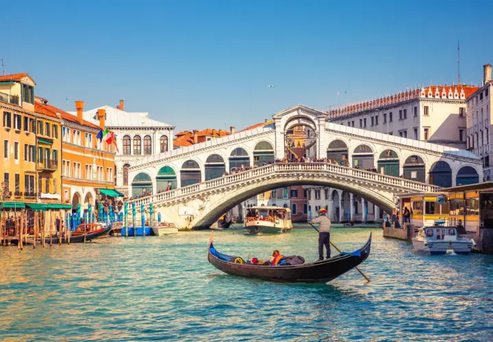 Gondole naviguant sur le Grand Canal, sous le célèbre pont du Rialto à Venise. L'eau turquoise reflète les façades colorées des bâtiments.