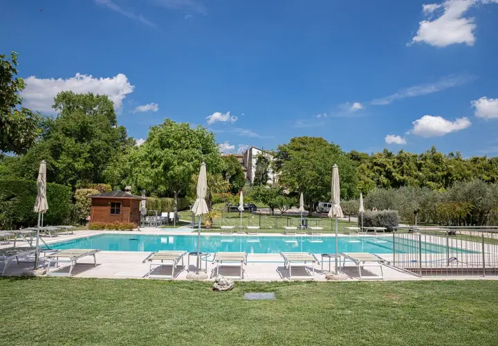 Piscine d'agritourisme au Lac de Garde, entourée de verdure, avec chaises longues et parasols sous un ciel bleu.