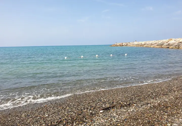 Plage de galets à Cecina Mare, avec des bouées blanches marquant la zone de baignade et une jetée rocheuse au loin.