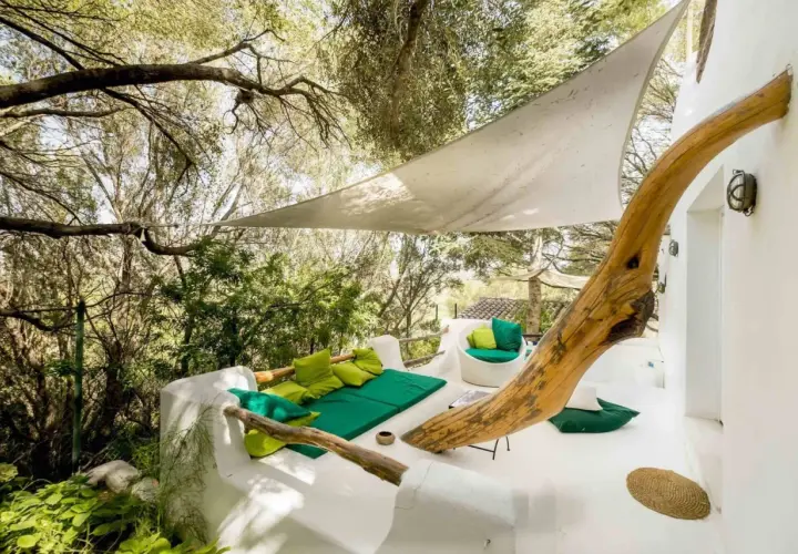 Terrasse du Hotel Su Gologone avec mobilier blanc et coussins verts, sous une voile d'ombrage et entourée de verdure.