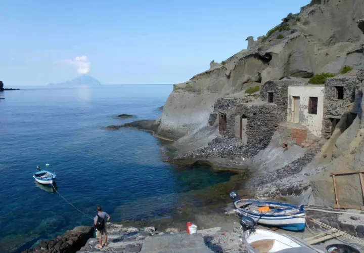 Sur l'île Salina, un homme amarre un bateau près de vieilles maisons de pêcheurs en pierre, avec une île volcanique au loin.