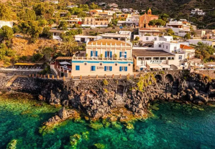 Vue d'un gîte en Sicile, perché sur une falaise rocheuse surplombant une mer turquoise. Bâtiments blancs et ocres, végétation luxuriante.