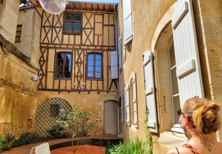 Cour intérieure avec colombages et volets blancs. Une femme regarde la scène, comme si elle découvrait un coin charmant de Fleurance, Italie.