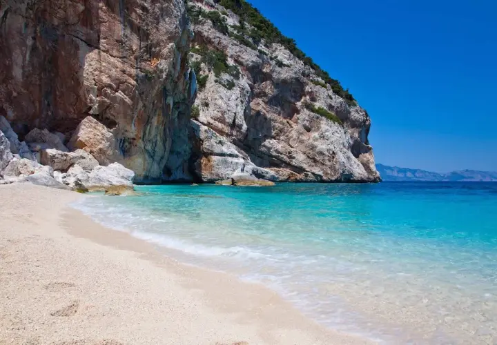Plage de sable fin bordée par une eau turquoise cristalline, avec une falaise imposante en arrière-plan. Un coin de paradis sur la côte smeralda sardaigne.