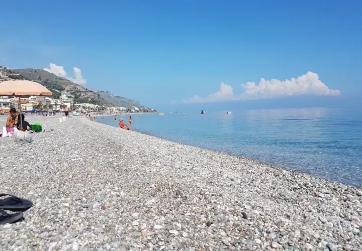 Plage de galets à Letojanni, Sicile. Des baigneurs profitent du soleil sous un parasol, tandis que la mer d'un bleu profond s'étend à l'horizon.