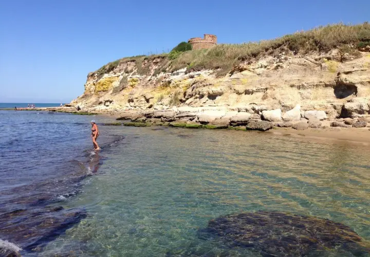 Une femme en bikini profite de la mer limpide près d'une falaise rocheuse à Anzio, avec une tour ancienne en arrière-plan.