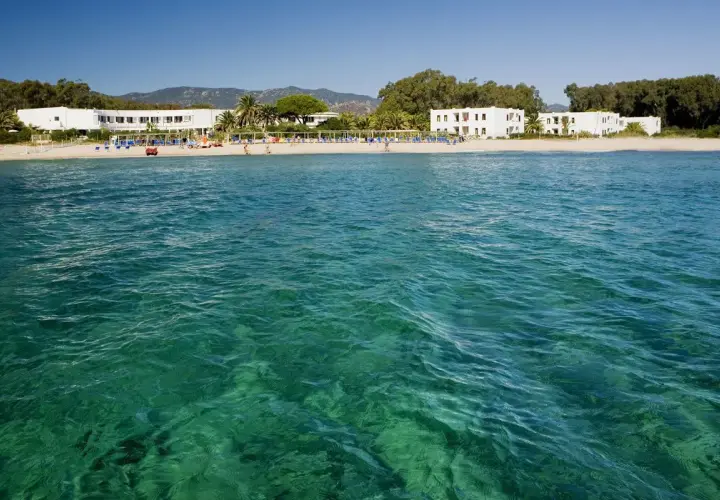 Eaux turquoise cristallines devant une plage de sable fin à Pula, Sardaigne, avec des hôtels blancs et des palmiers.