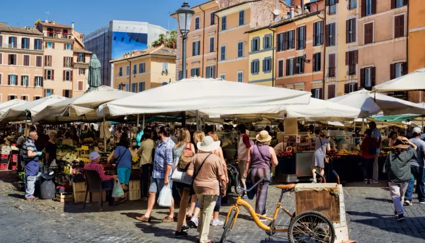 Animation d'un marché animé à Rome, avec des étals de fruits et légumes sous des parasols blancs. Une bicyclette jaune est garée près d'un chariot.