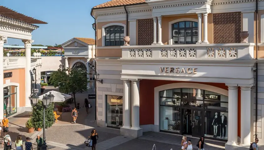 Boutiques de luxe, dont Versace, dans un centre commercial de style italien près de Rome. Des clients flânent sous le soleil.