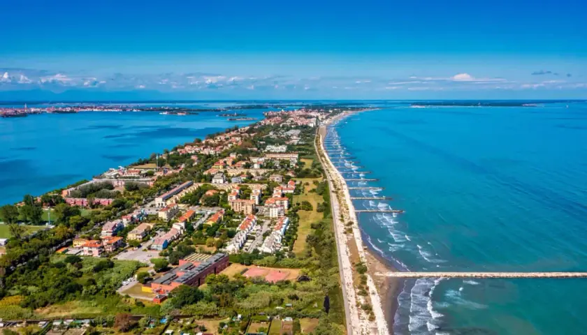 Vue aérienne du Lido di Venezia, une longue bande de terre bordée par la mer Adriatique et la lagune, avec des maisons, des arbres et des digues.