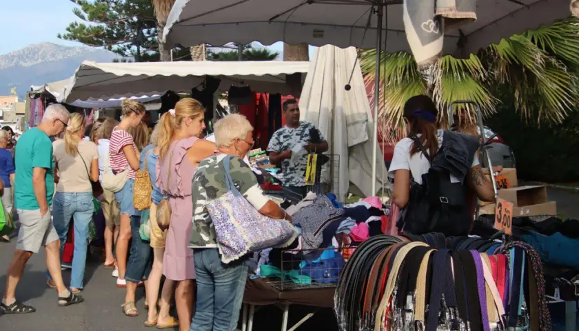 Jour de marché à Albenga : des gens flânent devant des étals de vêtements et d'accessoires, sous les palmiers.