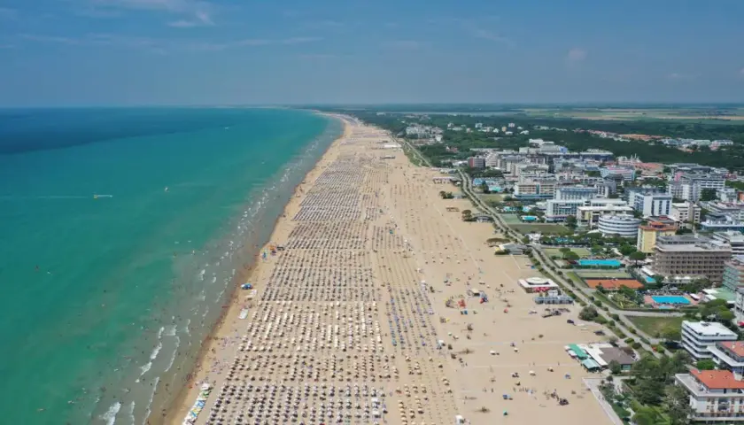 Plage bondée de parasols et de baigneurs à Bibione, Italie, avec la mer turquoise et des hôtels en arrière-plan.