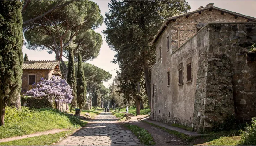 Chemin pavé bordé de cyprès et de pins, avec des maisons anciennes et des glycines en fleurs. Un air de via antica Roma.