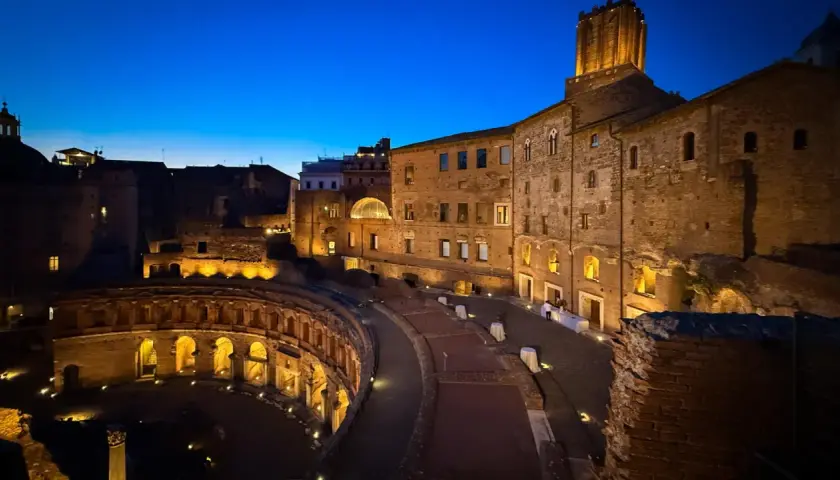Les marchés de Trajan s'illuminent dans la nuit romaine, vestiges antiques baignés d'une douce lumière dorée sous un ciel bleu profond.