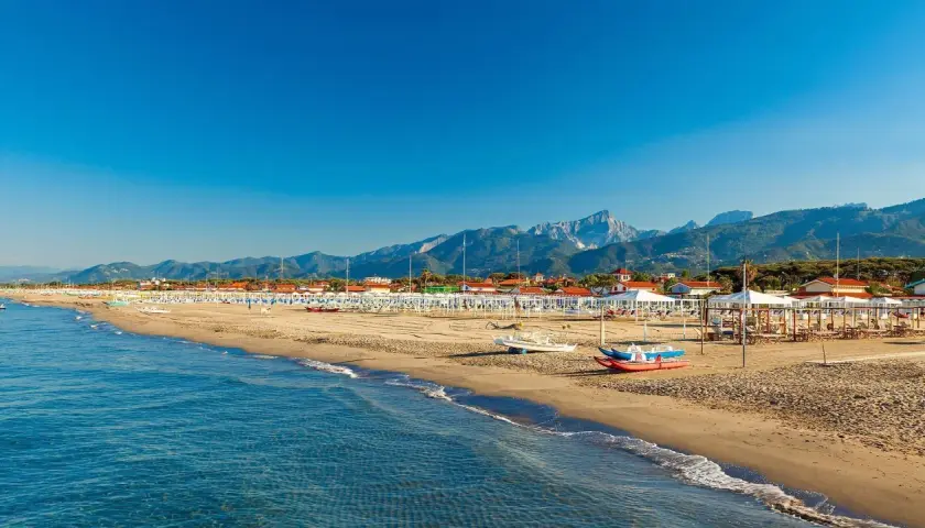 Plage de sable fin à Forte dei Marmi, Italie, avec des rangées de parasols blancs et des montagnes majestueuses en arrière-plan.