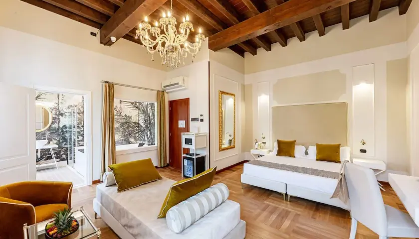 Suite luxueuse au **hotel Cariola Caprino Veronese** avec lit king-size, canapé, lustre et vue sur jardin tropical.