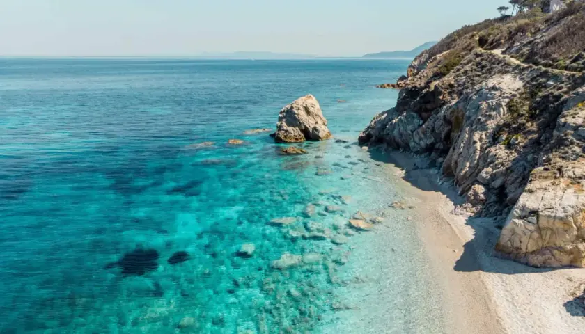 Plage de sable fin bordée par des eaux turquoise cristallines et des falaises rocheuses, typique des iles toscanes. Un rocher solitaire émerge de la mer.
