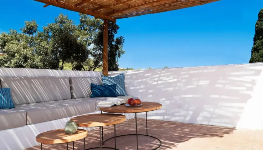 Terrasse ombragée d'un agriturismo en Sardaigne, avec canapé confortable, tables basses en bois et vue sur la végétation luxuriante sous un ciel bleu éclatant.
