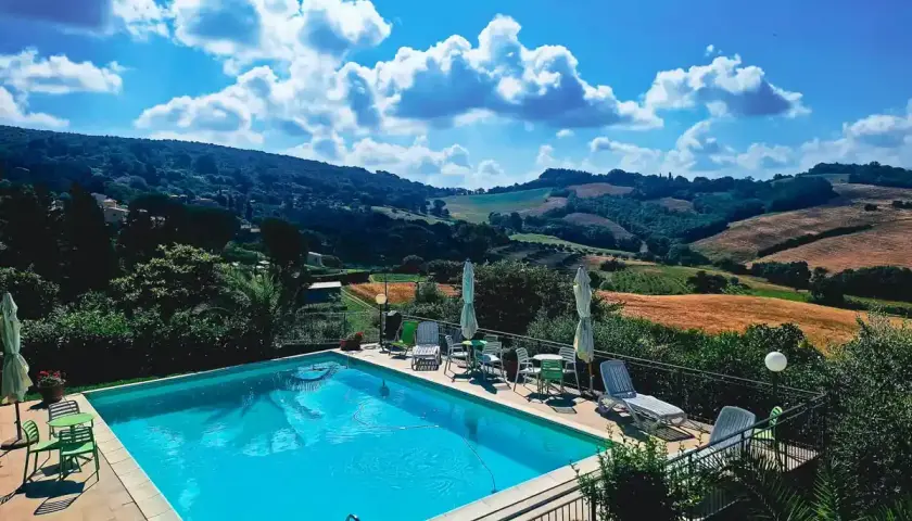Piscine étincelante devant une **ferme toscane** pittoresque, avec des collines ondulantes et un ciel bleu éclatant.