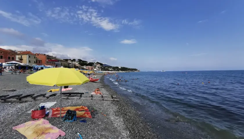 Plage de galets animée, avec des parasols, des serviettes et des baigneurs profitant du soleil. La mer bleue s'étend à l'horizon, avec des bateaux au loin. Un aperçu de la vie sur la côte ligure.