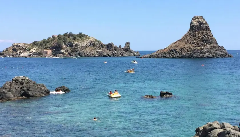 Vue sur la mer bleue et les rochers volcaniques d'Aci Trezza, Sicile. Des bateaux et des baigneurs profitent du soleil.
