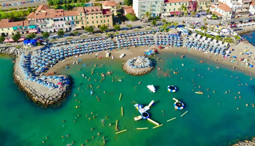 Plage animée à Arma di Taggia, Italie, avec des parasols bleus et blancs, des baigneurs dans la mer turquoise et des structures gonflables.