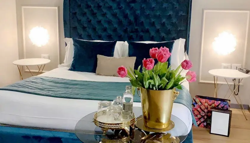 Chambre d'hôte en Lombardie : lit luxueux avec tête de lit capitonnée bleue, tulipes roses et tables de chevet élégantes.